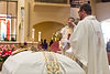 A priest in a white cassock blessing a casket covered in a white cloth with incense 